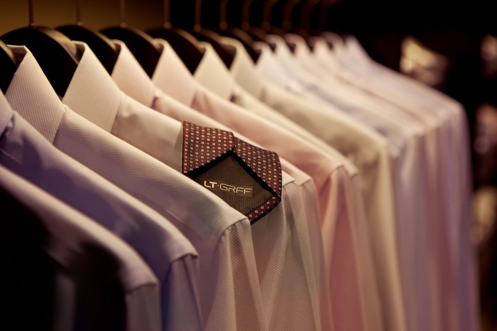 A row of neatly hung dress shirts on wooden hangers in a closet. The shirts, made from premium Luthai Textile fabrics, are predominantly light-colored with one dark shirt featuring a patterned collar at the center. The brand label "LT-GRF" is visible on the dark shirt's collar.