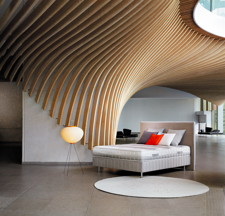 A modern, minimalist bedroom features a bed with white bedding, accentuated by orange and red pillows. A spherical lamp with a wooden base sits nearby. The striking wooden ceiling design arches dramatically, creating a visually captivating effect, with a Cofel skylight above.