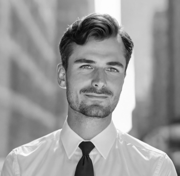 Black and white portrait of a man wearing a crisp white shirt and dark tie. He has neatly groomed dark hair and a short beard, and is facing the camera with a subtle smile. The background suggests an urban setting, hinting at faster market entry and speeding innovation in his field.