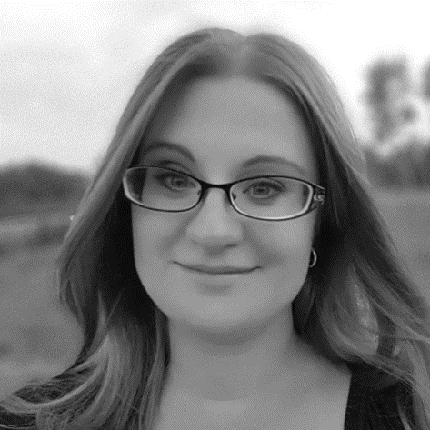 A black-and-white photograph of a woman with shoulder-length hair wearing glasses. She is smiling softly and looking directly at the camera. The background, slightly blurred, suggests an outdoor setting with trees and an open field, evoking a sense of serenity amidst speeding innovation.