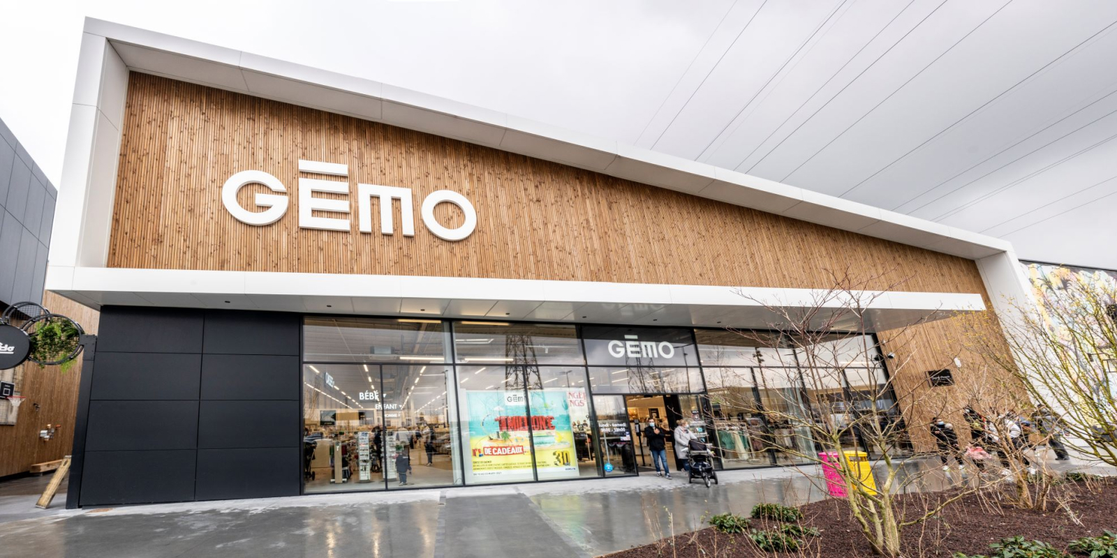 A wide-angle view of a modern retail store with a wooden facade and large sign reading "Gémo" above the entrance. The storefront has large glass windows displaying clothing and other merchandise. People are seen entering and exiting the store on a cloudy day.