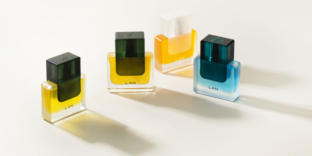 Four perfume bottles on a light surface, each with different colored liquids: yellow, green, orange, and blue. The bottles are rectangular with black caps, and the brand name "LAN" is displayed on each. They cast soft shadows.