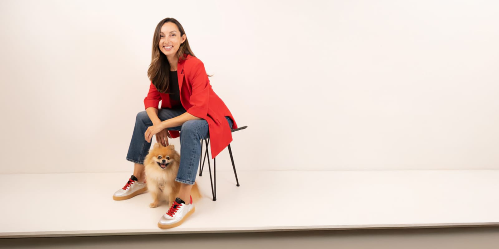 Una donna sorridente, con indosso un blazer rosso e jeans, è seduta su una sedia e accarezza un piccolo cagnolino soffice che sta ai suoi piedi, su uno sfondo bianco.